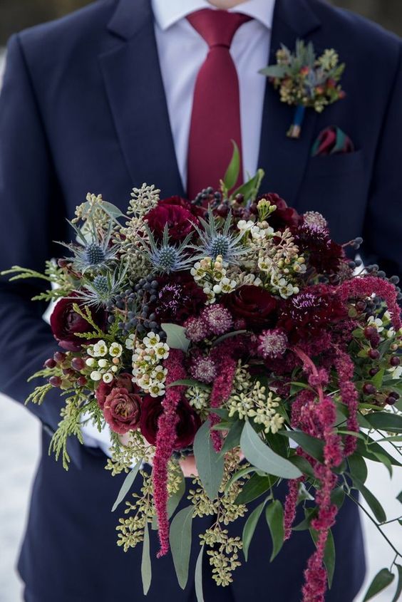 a moody wedding bouquet of deep burgundy roses and other blooms, waxflowers, thistles, greenery and amaranthus for fall or winter