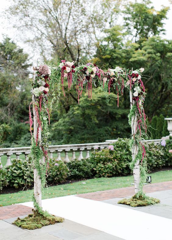 a rustic wedding arch covered with greenery, white and burgundy blooms and amaranthus and moss is a cool idea for a rustic or woodland wedding