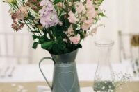 a rustic wedding centerpiece of a metal jug and pastel and purple blooms, baby’s breath in tin candles and a wood slice as a stand