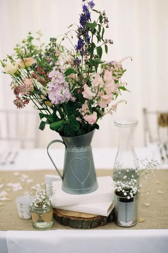 a rustic wedding centerpiece of a metal jug and pastel and purple blooms, baby's breath in tin candles and a wood slice as a stand