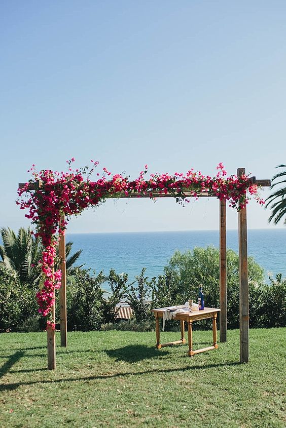 a simple wooden wedding arch with bougainvillea and a sea view is a cool and bold idea for a bright summer wedding