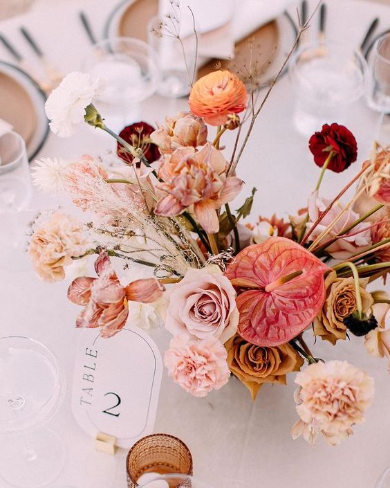 a sunset-colored wedding centerpiece of blush roses, carnations, anthuriums, grasses and ranunculus is amazing