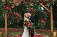 a super creative farmhouse wedding arch with burgundy, blush and red blooms, greeneery, amaranthus built over a large stone