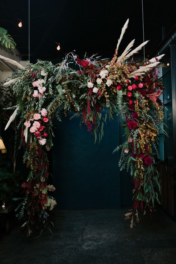 a super lush and moody wedding arch with greenery, blush, fuchsia, burgundy blooms, foliage, pampas grass and amaranthus for a fall boho wedding