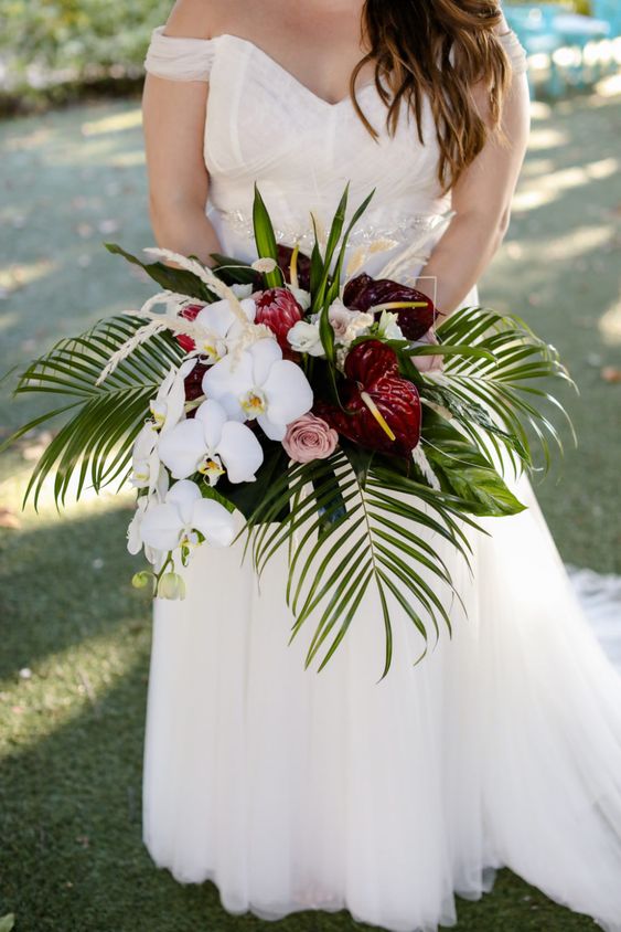 a tropical wedding bouquet of white orchids, blush roses, king protea and burgundy anthurium and leaves