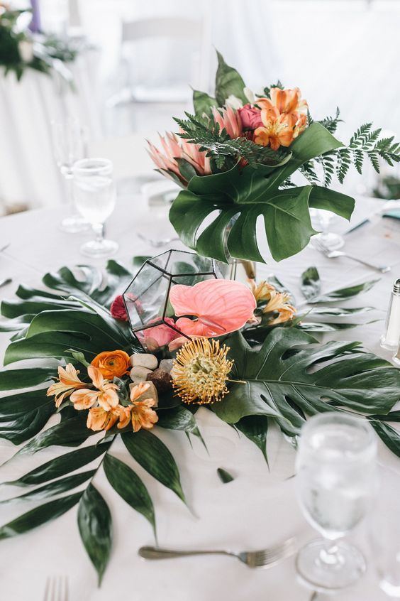 a tropical wedding centerpiece of tropical leaves, pincushions proteas, yellow ranunculus, anthuriums in terrariums