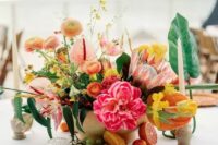 a vibrant wedding centerpiece of coral ponies, king proteas, peachy ranunculus, greenery and anthuriums, fruits on the table