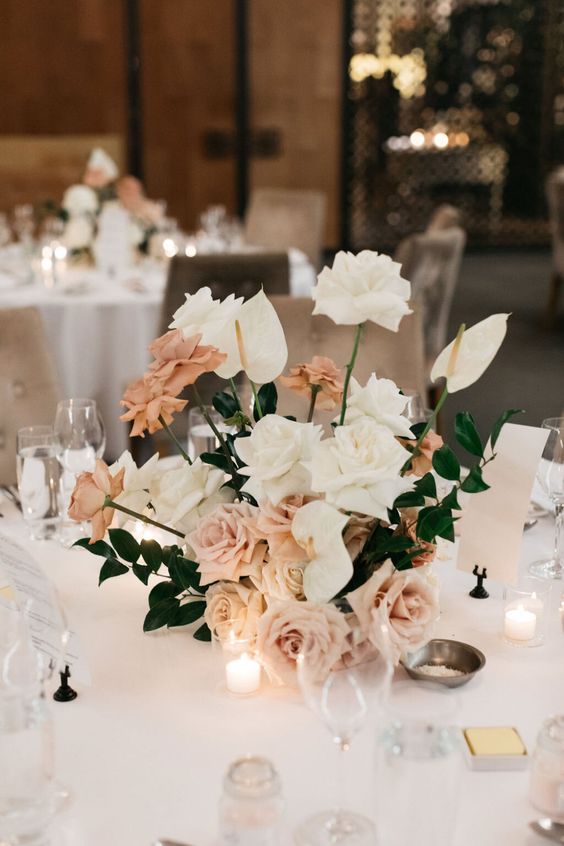 an exquisite wedding centerpiece of blush and white roses, greenery and anthuriums plus candles around is amazing