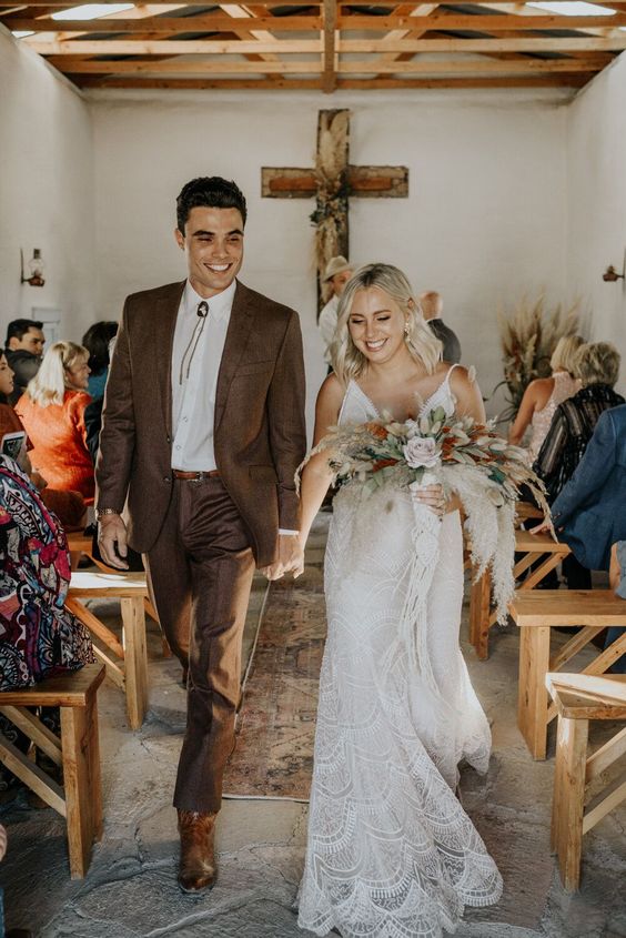 a boho groom's outfit with a brown pantsuit, a white shirt, brown shoes and a bolo tie is cool for the fall