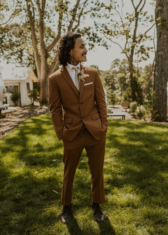 a brown pantsuit, a white shirt, a tan tie, black shoes and a dried flower boutonniere for a boho groom