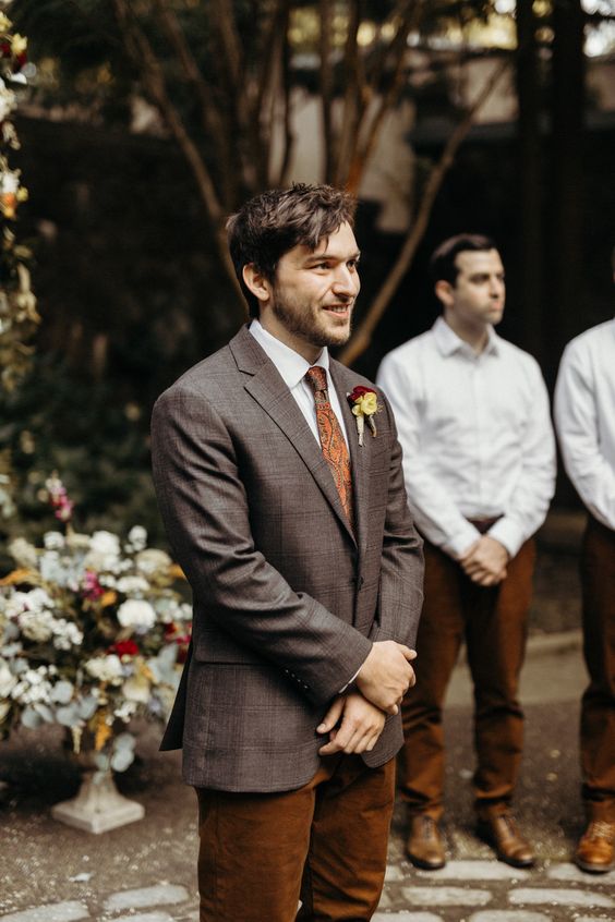 a catchy boho fall groom's look with a white shirt, rust trousers, a brown blazer, a rust tie and a bold boutonniere
