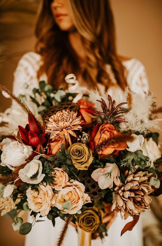 a beautiful and bold fall wedding bouquet of peachy and dark roses, white blooms, rust roses and chrysanthemums, greenery and feathers