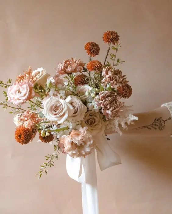 a beautiful fall wedding bouquet of blush roses, pink chrysanthemums, marigolds, greenery and long ribbon is chic