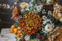 a bold and catchy wedding centerpiece of burgundy and peachy chrysanthemums, greenery, seed pods, daisies, citrus