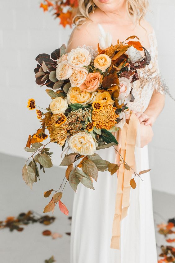 a bold fall wedding bouquet of peachy roses, blush and yellow ranunculus, chrysanthemums, bold foliage and bright leaves