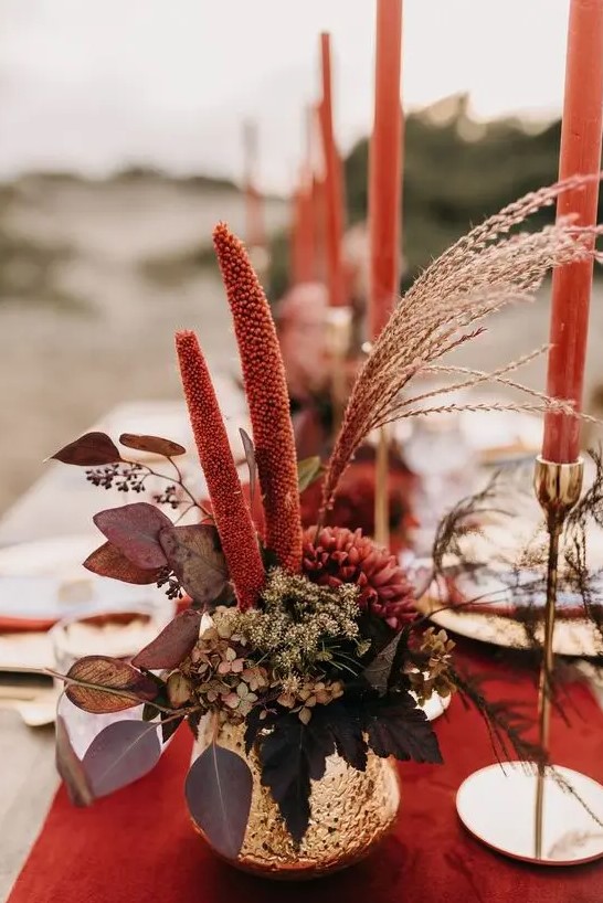 a bold fall wedding centerpiece in a gold vase, with burgundy blooms, greenery and dark foliage plus red candles