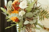 a bold fall wedding centerpiece of white and burgundy blooms, orange chrysanthemums, greenery and seed pods