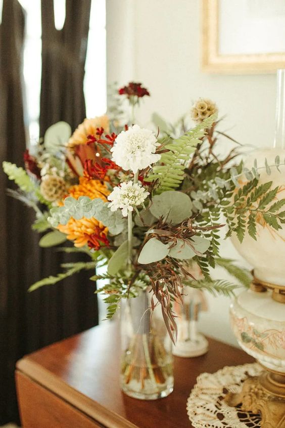 a bold fall wedding centerpiece of white and burgundy blooms, orange chrysanthemums, greenery and seed pods