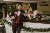 a catchy fall groom’s outfit with a burgundy suit, a red waistcoat, a white shirt, brown shoes and a printed bow tie