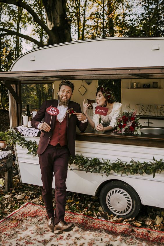 a catchy fall groom's outfit with a burgundy suit, a red waistcoat, a white shirt, brown shoes and a printed bow tie