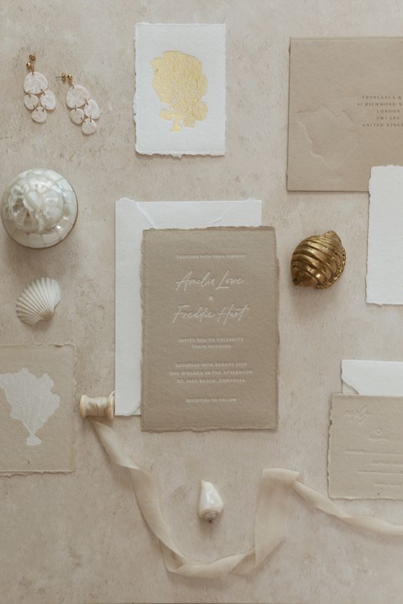 a chic coastal wedding invitation suite with a white envelope, beige cards, a godl coral print and a raw edge
