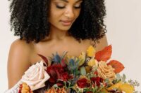 a colorful fall wedding bouquet of red and rust roses, rust chrysanthemums, thistles, greenery and bold fall foliage is wow
