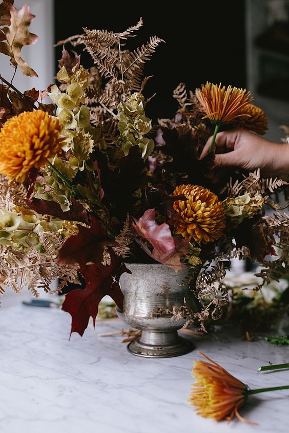 a moody fall wedding centerpiece of mustard mums, dark foliage, some yellow blooms and dried leaves is amazing