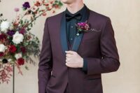 a moody groom’s look with a plum-colored tuxedo with black lapels, a black shirt and a bow tie, a bold boutonniere