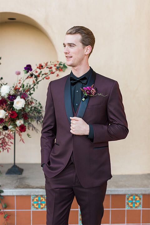 a moody groom's look with a plum-colored tuxedo with black lapels, a black shirt and a bow tie, a bold boutonniere