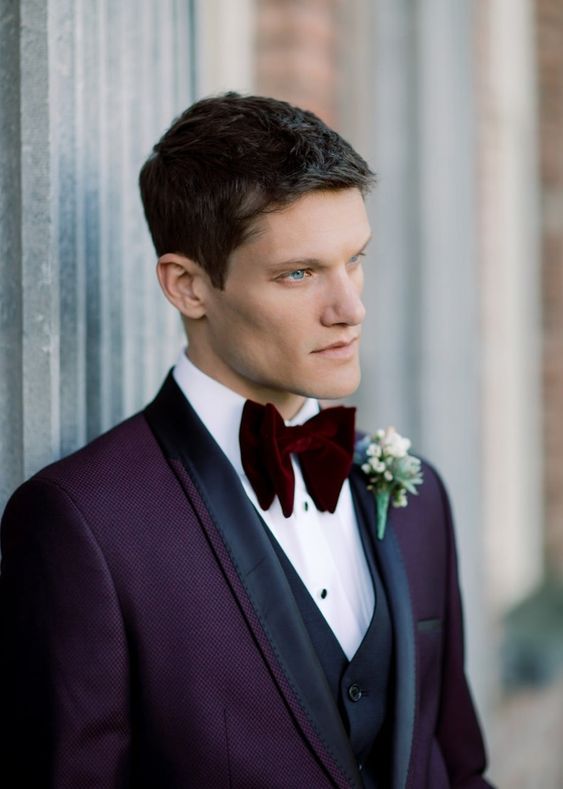 a sophisticated groom's look with a purple tux with black lapels, a white shirt, a black waistcoat and a burgundy velvet bow tie