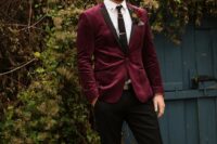 a stylish fall groom’s look with a burgundy velvet blazer with black lapels, black pants, black shoes, a white shirt and a black tie plus chains