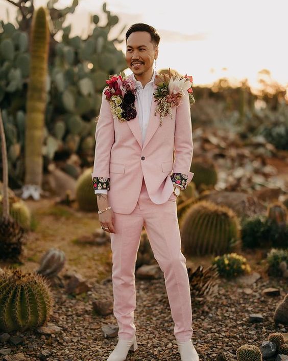 a unique boho groom's look with a blush pantsuit, a white shirt, creamy boots, flower adorned shoulders is wow