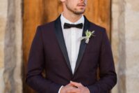 an elegant purple groom’s look with a tuxedo, black lapels, a blakc bow tie and a flower boutonniere