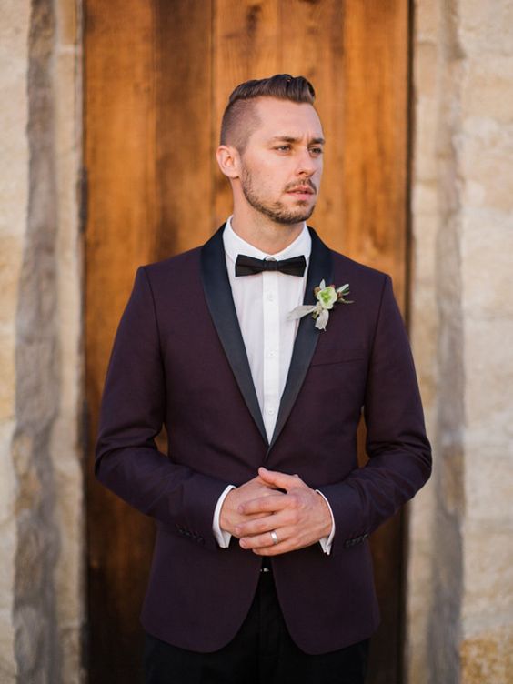 an elegant purple groom's look with a tuxedo, black lapels, a blakc bow tie and a flower boutonniere
