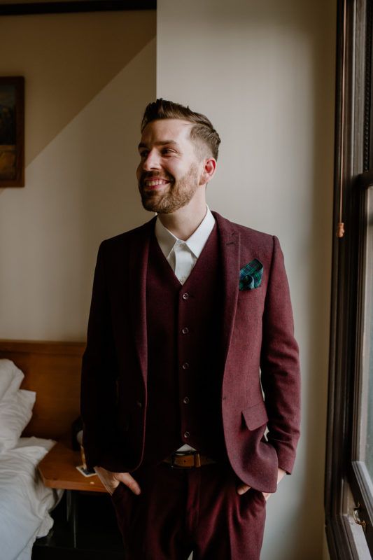 an elegant vintage-inspired groom's look with a burgundy three-piece pantsuit, a white shirt, an amber leather belt