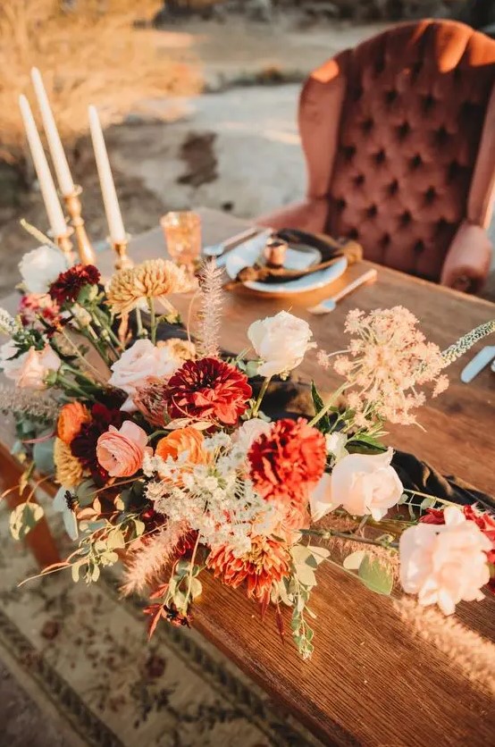 an exquisite boho fall wedding centerpiece of blush roses, burgundy mums and ranunculus, greenery and grasses is amazing for a fall celebration