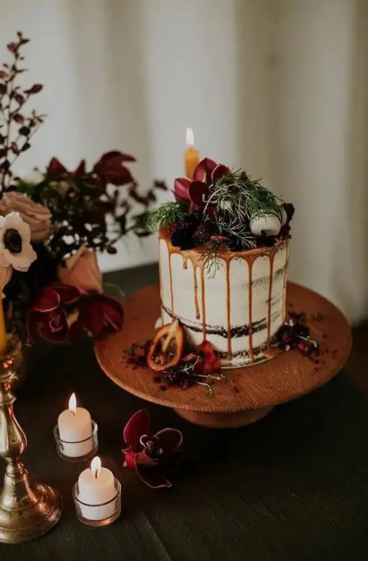 a chocolate naked wedding cake with caramel drip, fresh berries and dark blooms, macarons and greenery
