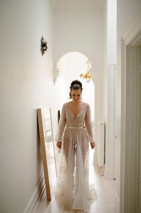 a neutral sheer wedding gown with embellishments all over it, a V-neckline, long sleeves and a bodysuit underneath