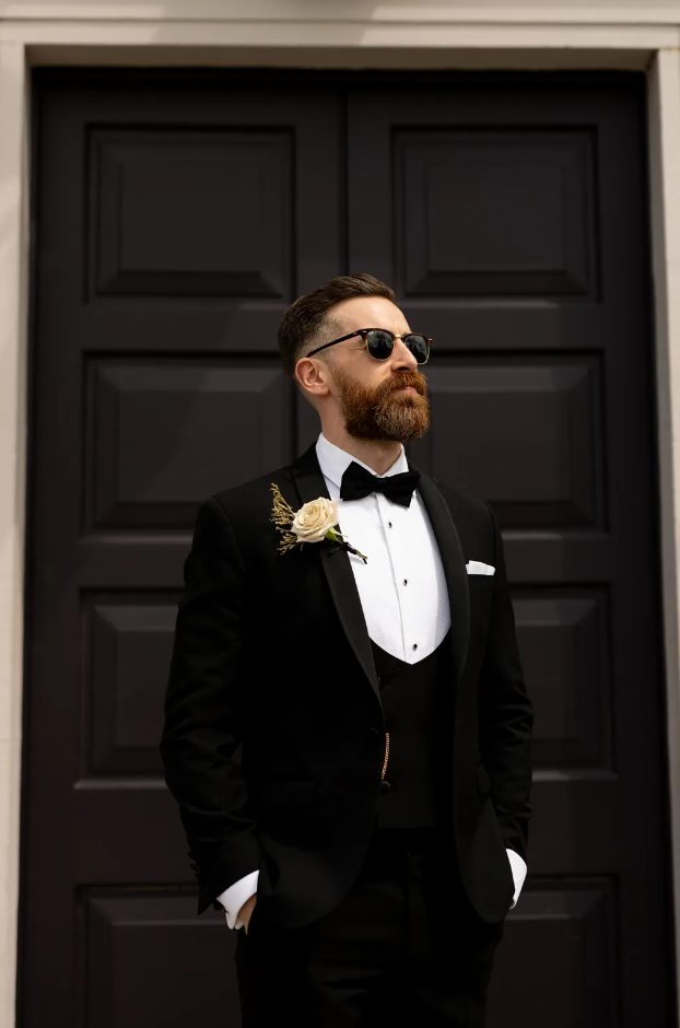 a stylish and refined black tux with a white shirt with black buttons, a chain, a hndkerchief and a white rose boutonniere