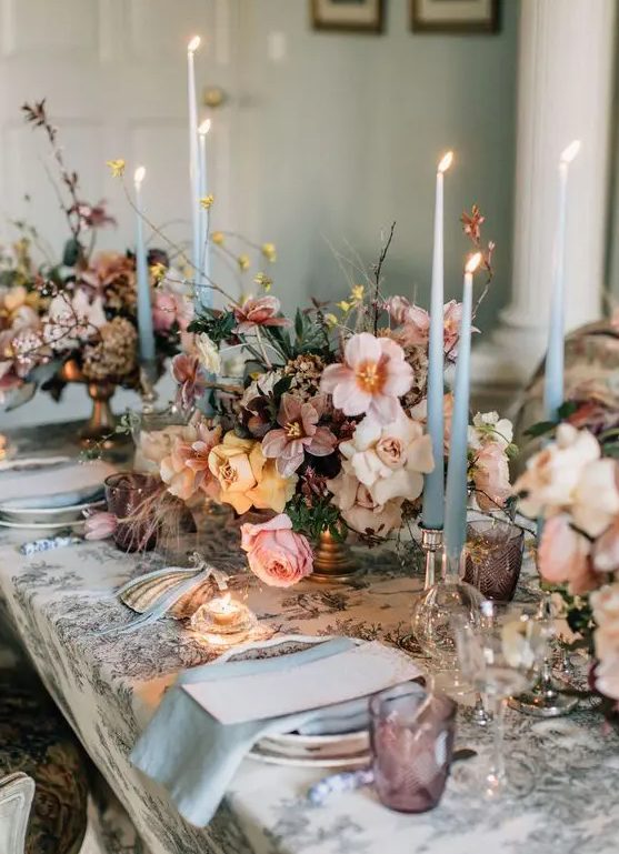 a fantastic wedding tablescape with a printed tablecloth, lush blush, peachy and mauve blooms, thin and tall blue candles and napkins