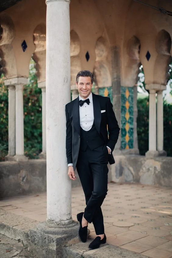 a stylish and timeless black tux with a white shirt and black buttons, black velvet loafers and a black bow tie