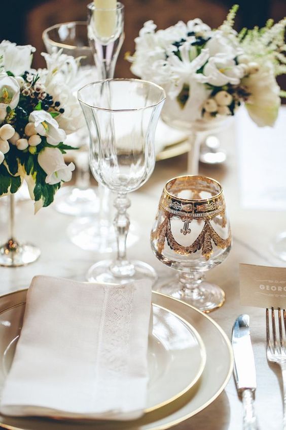 a chic white wedding tablescape with gold rimmed plates, white blooms, some gold-rimmed glasses and a vintage patterned one