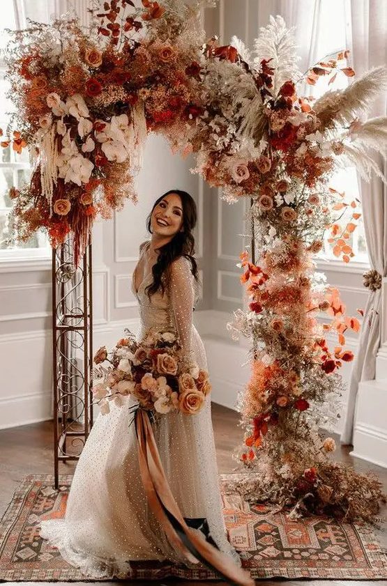 an incredible bright boho fall wedding arch with rust-colored and pink and blush roses, white orchids, pampas grass, rust-colored fall foliage just excites