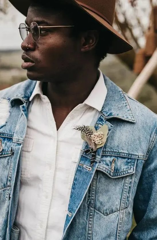 a cool relaxed groom's outfit with a white shirt, a blue denim jacket with a boutonniere and a brown hat is great for the fall