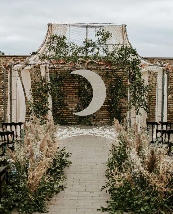 a beautiful boho celestial wedding ceremony space with a shiny half moon, greenery, textiles, greenery and pampas grass lining up the aisle