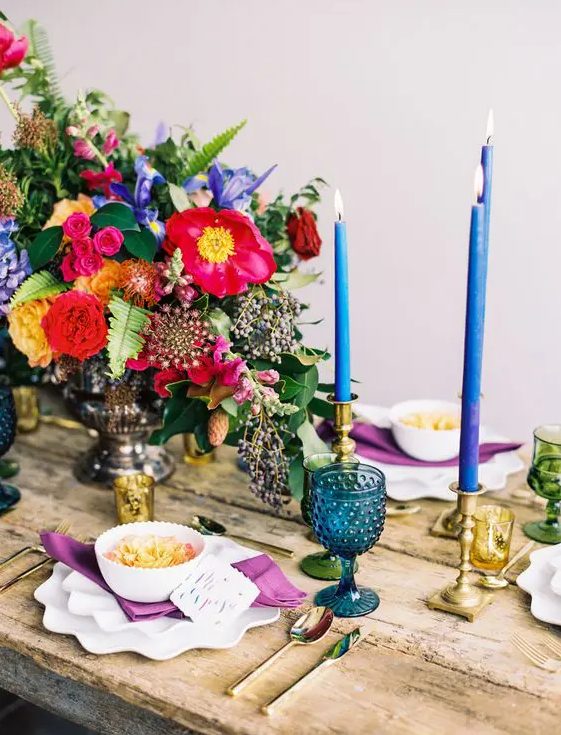 a bold Spanish-inspired wedding tablescape with hot pink, yellow and blue blooms, blue candles and glasses and purple napkins