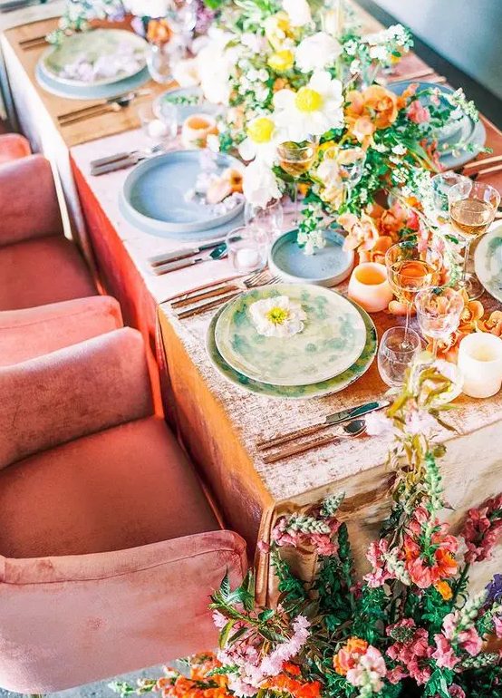 a bright wedding table setting with yellow, orange and white blooms, a peachy tablecloth and colorful plates, peachy candleholders and pink and orange blooms on the floor
