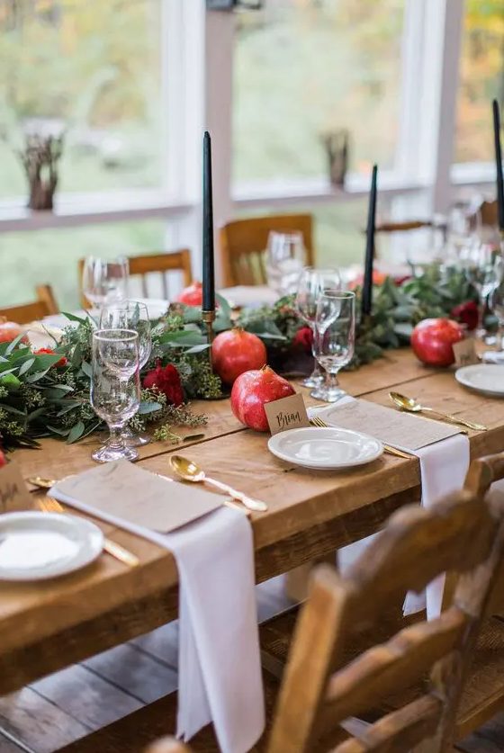 an exquisite fall wedding table setting with a greenery and burgundy bloom runner, pomegranates and black candles, gold cutlery