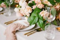a beautiful peach and cream wedding table with peachy and white blooms, greenery and blush napkins plus gold cutlery