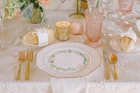 a peach and cream wedding table setting with peachy and white blooms and greenery, peachy glasses, gold cutlery and gold candleholders
